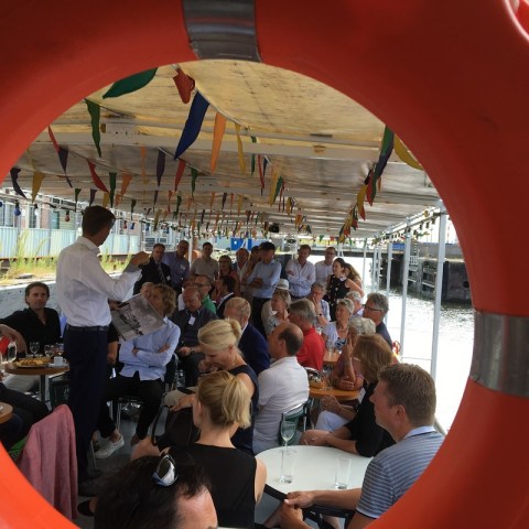 View through a lifebelt onto a man talking to many people sitting and standing on the boat