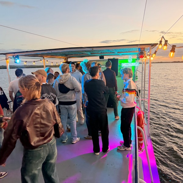 People gathered on a boat deck at sunset, with colorful lights and water in the background.