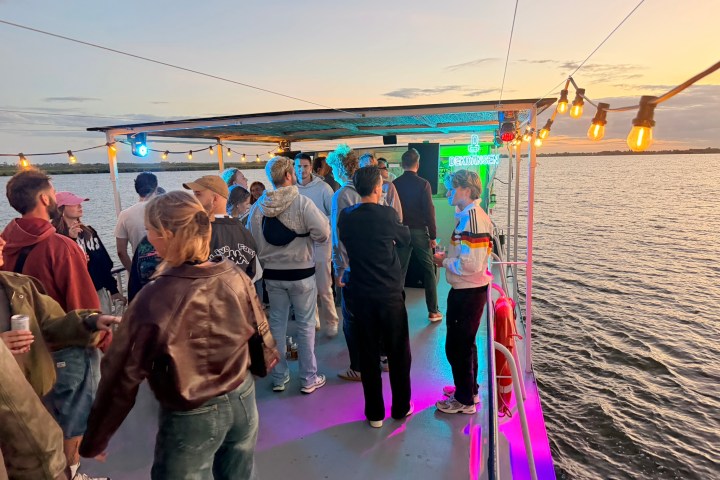 People gathered on a boat deck at sunset, with colorful lights and water in the background.