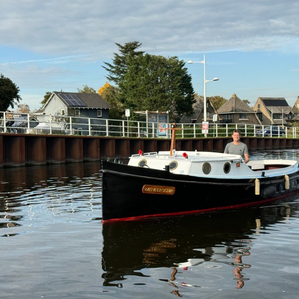 A man steers a black and white boat on a canal with houses in the background.