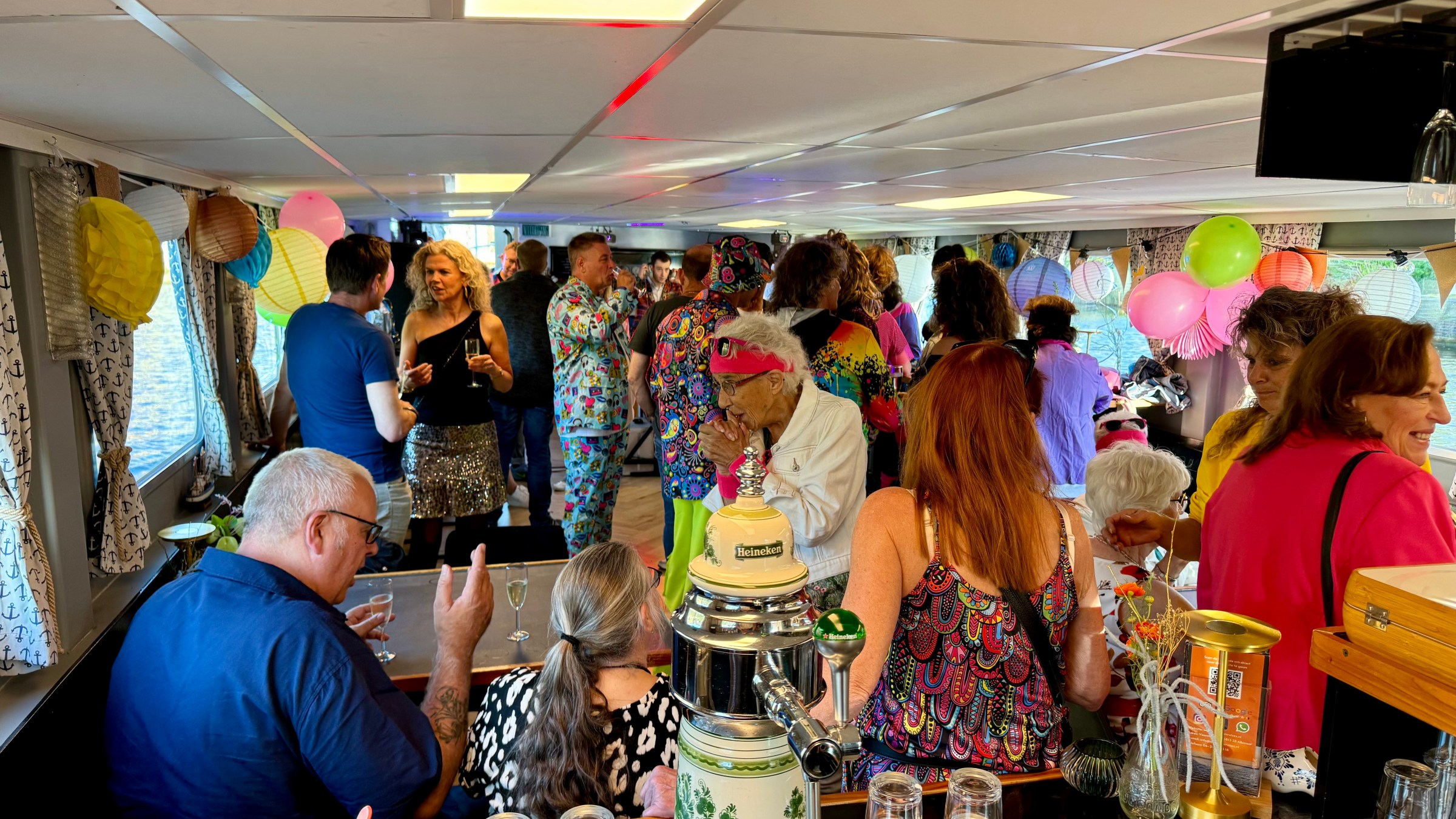 Crowded boat party with colorful clothes, balloons, and drinks; people mingling indoors.