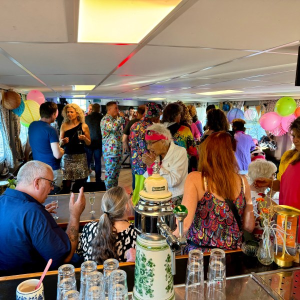 Crowded boat party with colorful clothes, balloons, and drinks; people mingling indoors.
