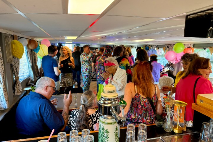 Crowded boat party with colorful clothes, balloons, and drinks; people mingling indoors.