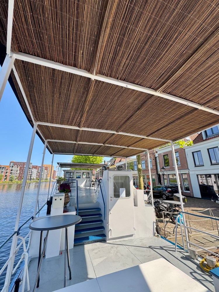 Boat deck with bamboo roof, moored by a canal with buildings in the background on a sunny day.
