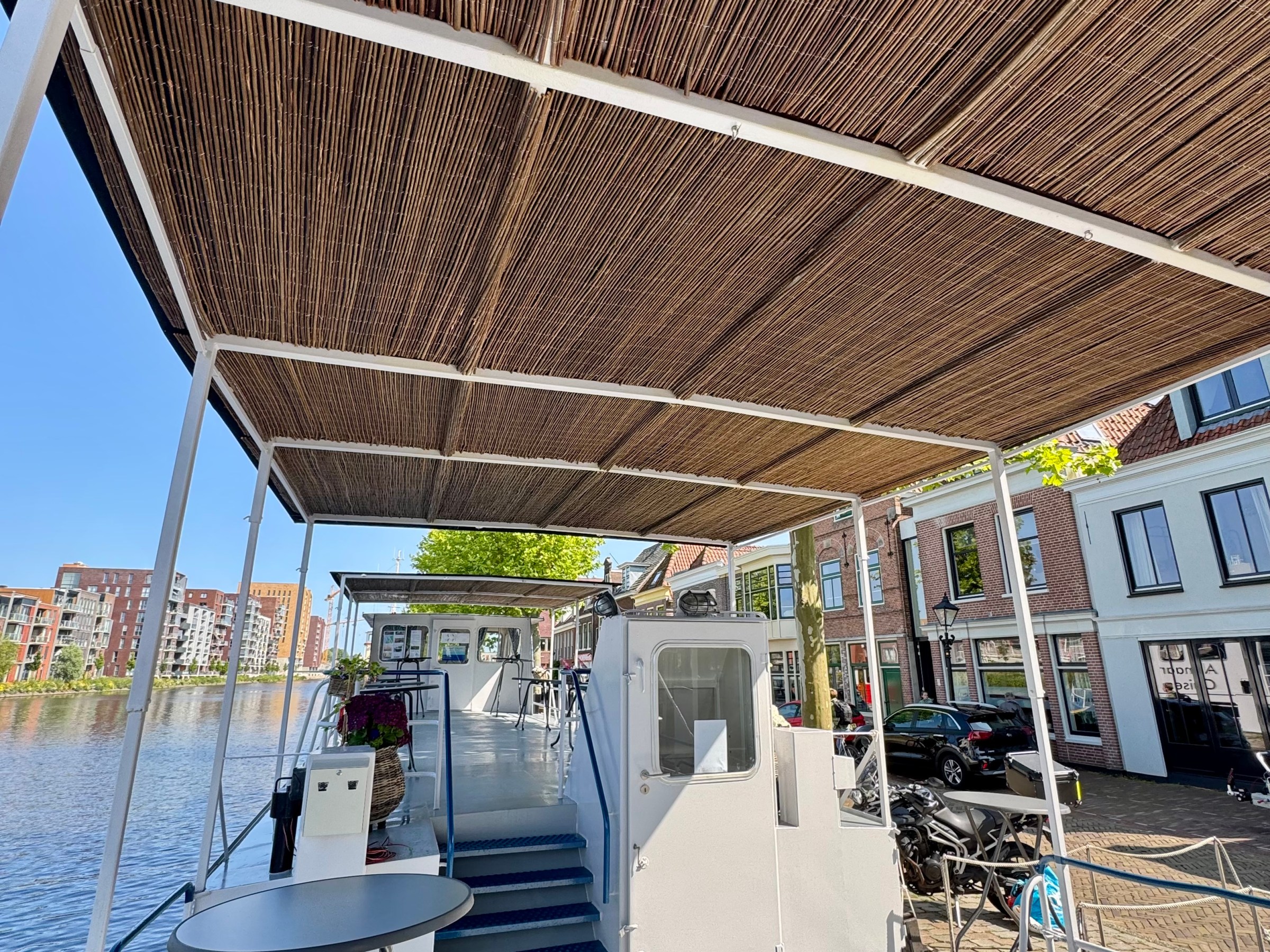 Boat deck with bamboo roof, moored by a canal with buildings in the background on a sunny day.
