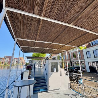 Boat deck with bamboo roof, moored by a canal with buildings in the background on a sunny day.