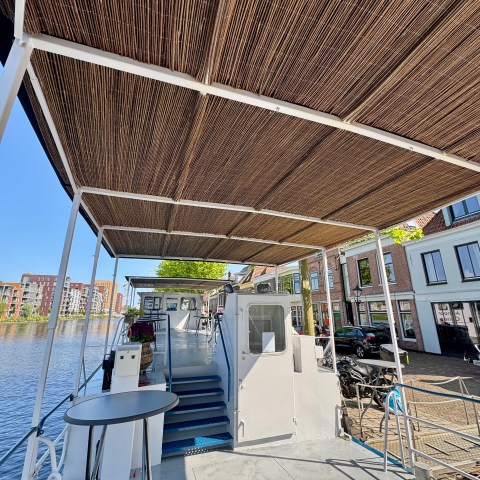 Boat deck with bamboo roof, moored by a canal with buildings in the background on a sunny day.