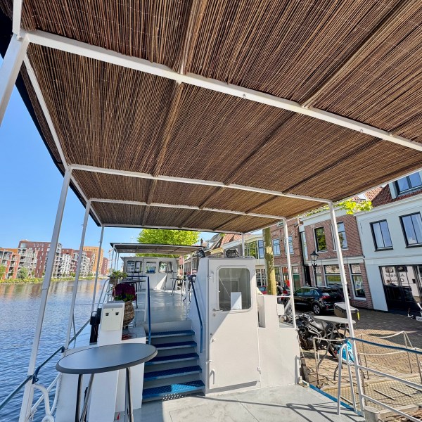 Boat deck with bamboo roof, moored by a canal with buildings in the background on a sunny day.