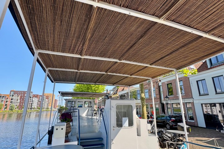 Boat deck with bamboo roof, moored by a canal with buildings in the background on a sunny day.