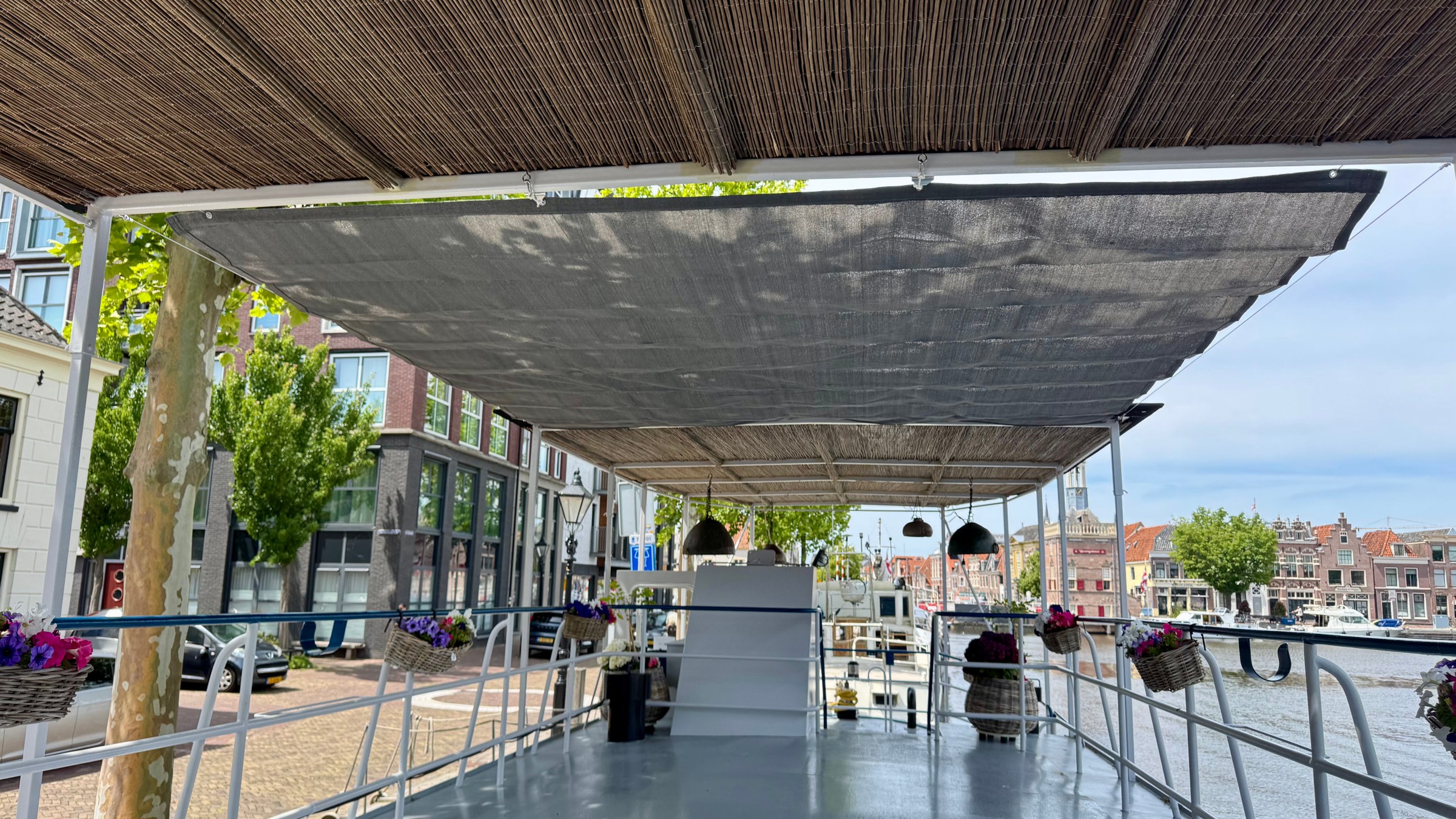 Boat deck with wicker roof and hanging baskets, docked near a canal and buildings.