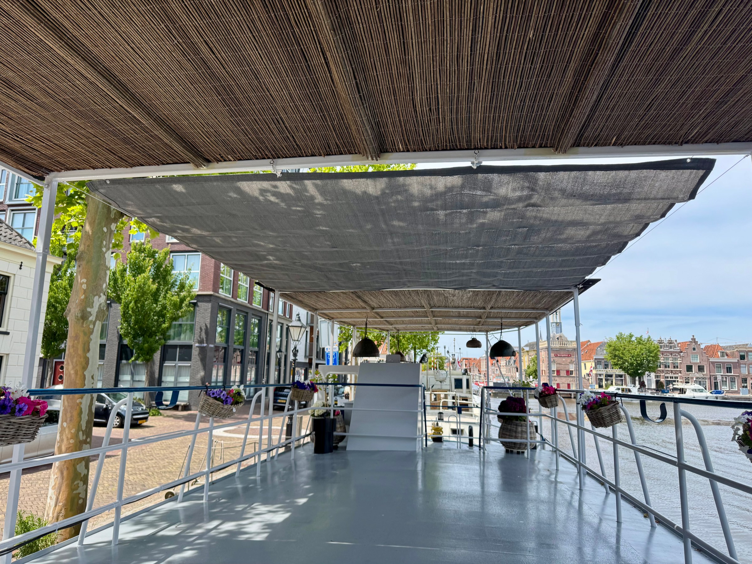 Boat deck with wicker roof and hanging baskets, docked near a canal and buildings.