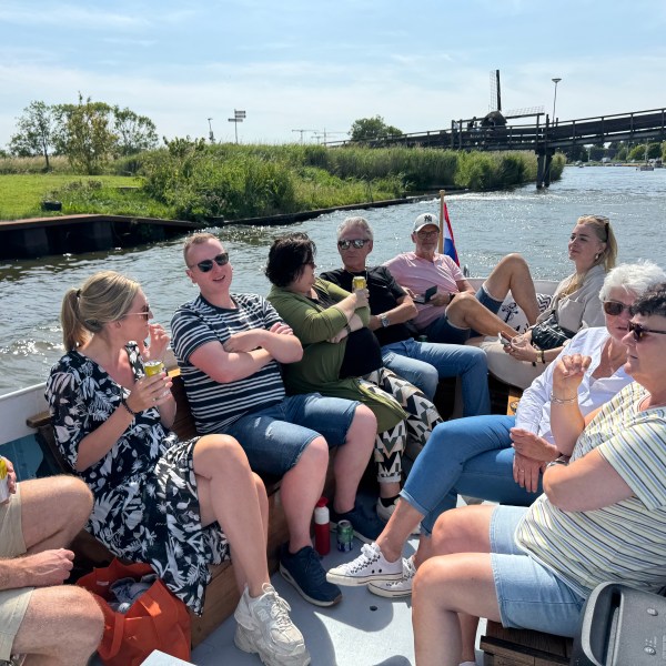Group of people enjoying a boat ride on a sunny day, seated and talking, with a windmill visible in the background.