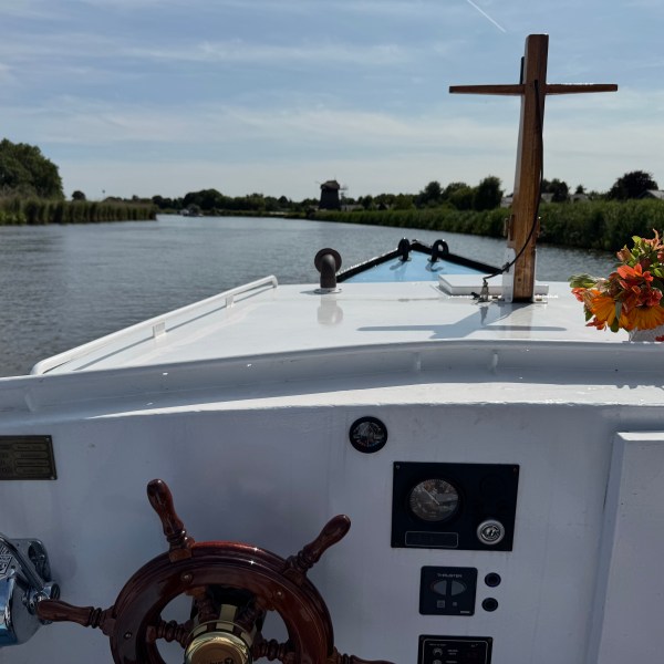 View from boat's helm with steering wheel, cross, and flowers on river.