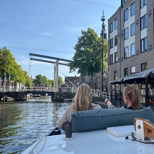 Two people on a boat in a canal with buildings and a bridge in the background.
