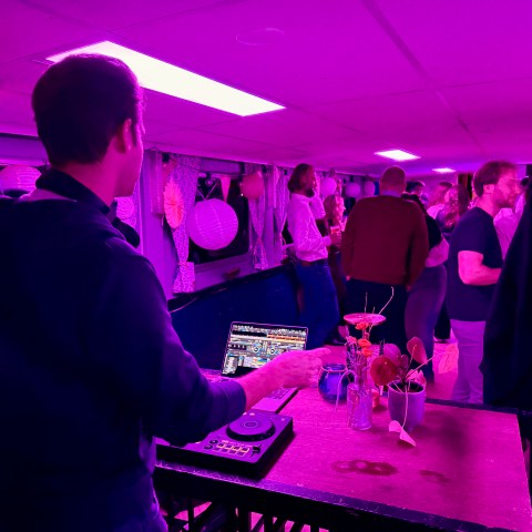 DJ playing music at a pink-lit party on a boat with people dancing.