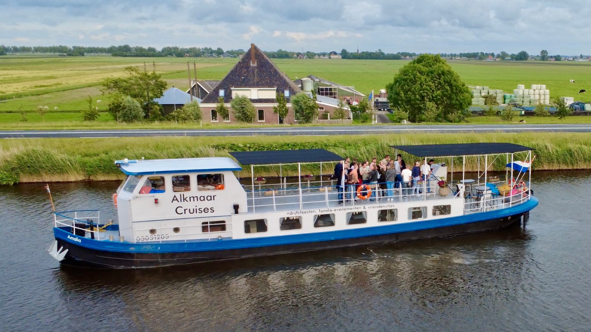 Tour boat 'Alkmaar Cruises' on a canal with passengers, farmland and rustic buildings in the background.