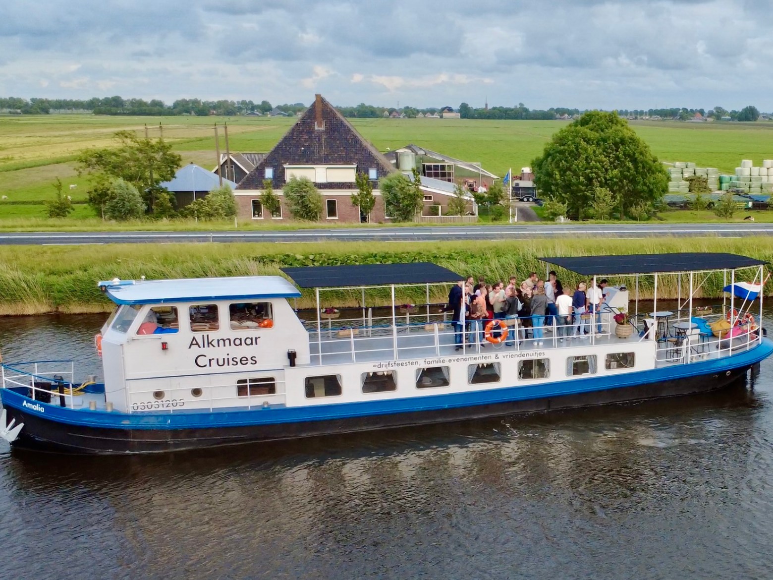 Tour boat 'Alkmaar Cruises' on a canal with passengers, farmland and rustic buildings in the background.