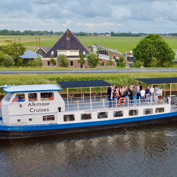 Tour boat 'Alkmaar Cruises' on a canal with passengers, farmland and rustic buildings in the background.