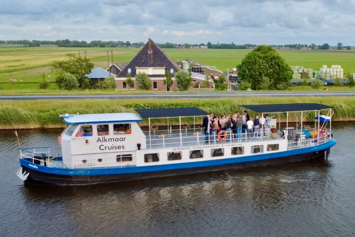 Tour boat 'Alkmaar Cruises' on a canal with passengers, farmland and rustic buildings in the background.