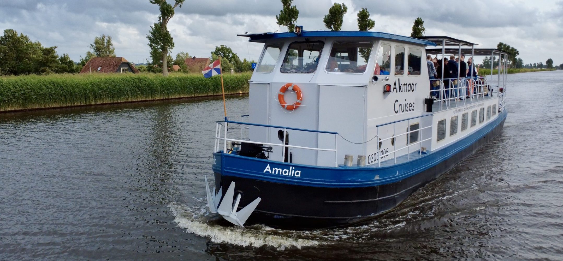 A blue and white passenger boat on a river with trees and a cloudy sky.