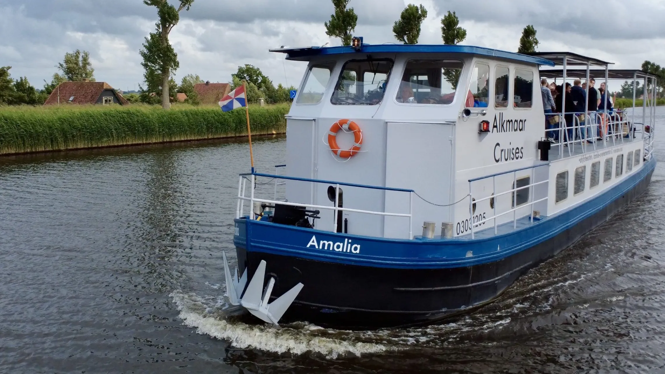 A blue and white passenger boat on a river with trees and a cloudy sky.